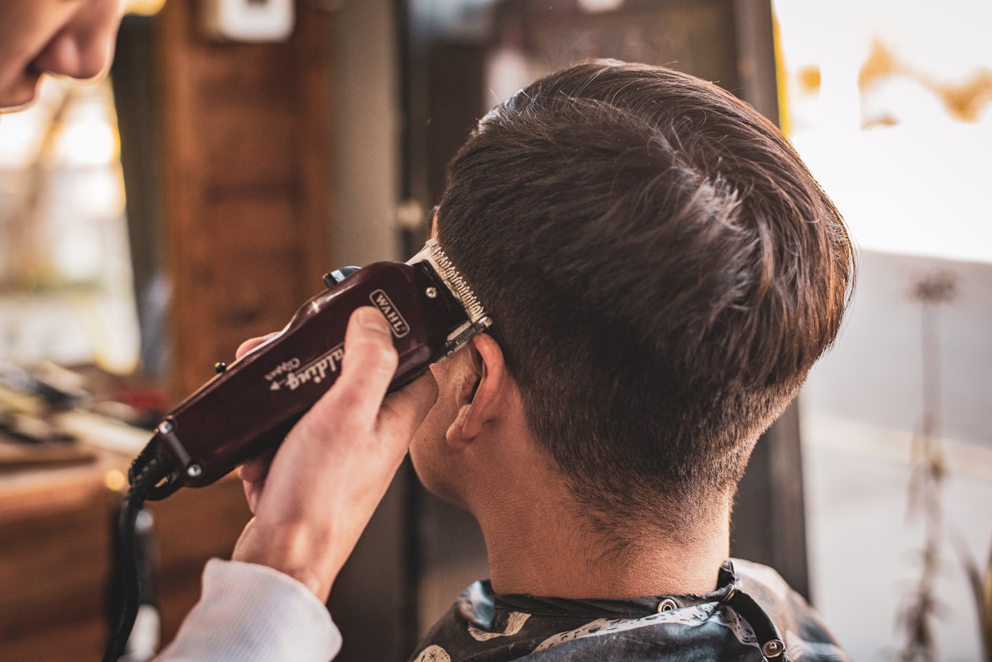 Barber trims customer's dark brown hair with electric clippers.